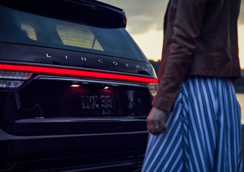 A person is shown near the rear of a 2024 Lincoln Aviator® SUV as the Lincoln Embrace illuminates the rear lights | Allan Vigil Lincoln, Inc. in Morrow GA
