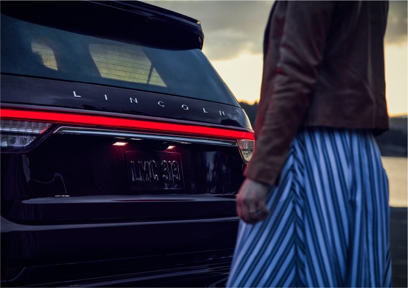 A person is shown near the rear of a 2023 Lincoln Aviator® SUV as the Lincoln Embrace illuminates the rear lights | Allan Vigil Lincoln, Inc. in Morrow GA