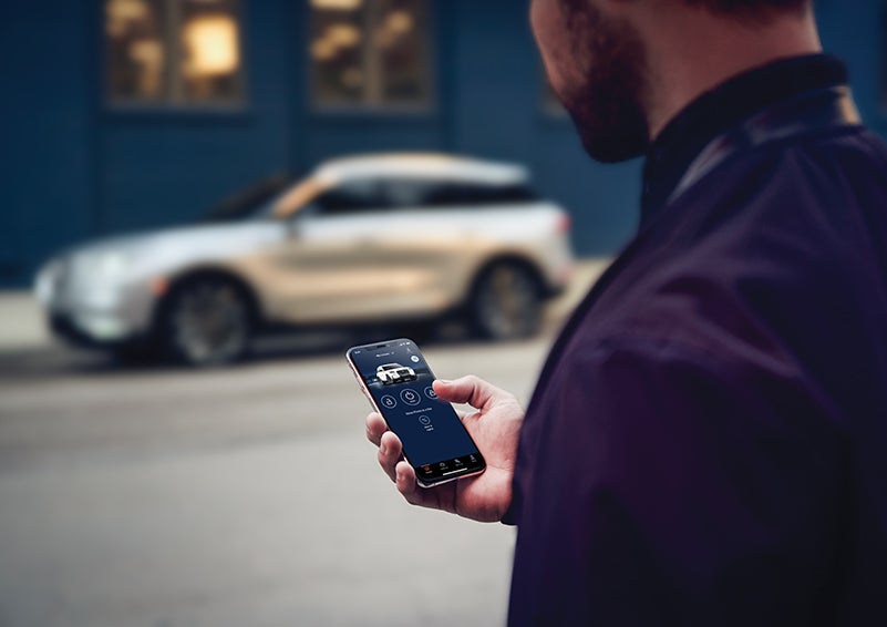 A person is shown interacting with a smartphone to connect to a Lincoln vehicle across the street. | Allan Vigil Lincoln, Inc. in Morrow GA